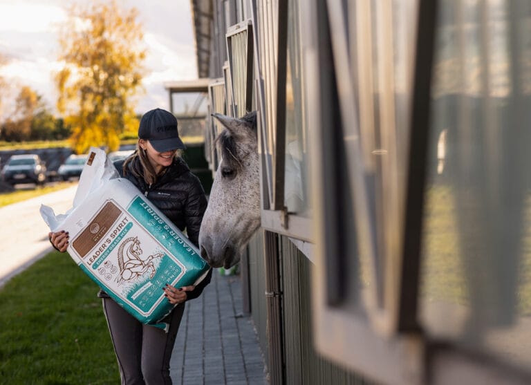 Uśmiechnięta kobieta trzymająca worek sianokiszonki Leader's Spirit Energy Grass Mix marki My Land 20 kg, stojąca przy boksie stajni. Siwy koń wyciąga łeb z boksu w jej stronę. W tle jesienny krajobraz.